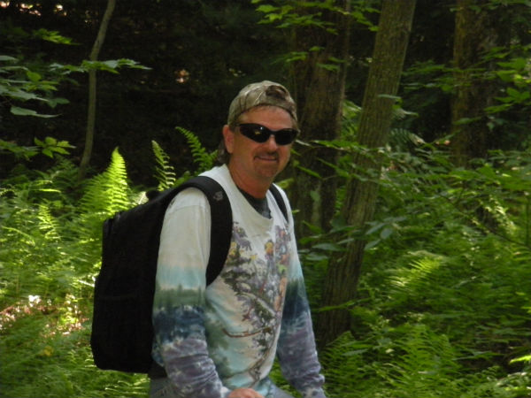 A man with sunglasses and backpack walking through the woods.