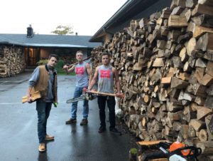 3 men with woodcutting tools next to stack of firewood