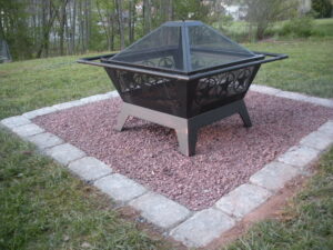 closeup of black portable firepit sitting on red stone base surrounded by white pavers in grass of backyard