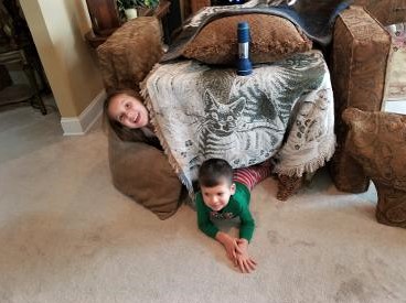 2 young kids on carpet floor inside a fort they stacked with furniture cushions and blankets