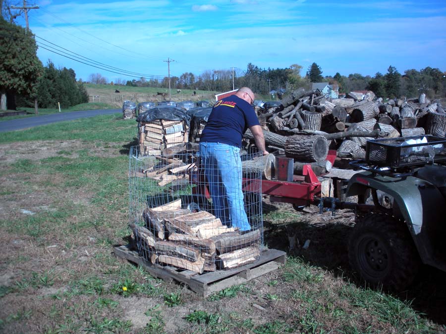 man splitting firewood with log splitter in field and loading wood onto pallets with metal fencing holding wood pile in tact