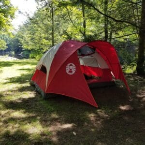 Large red and white camping tent pitched under trees along a mountain stream