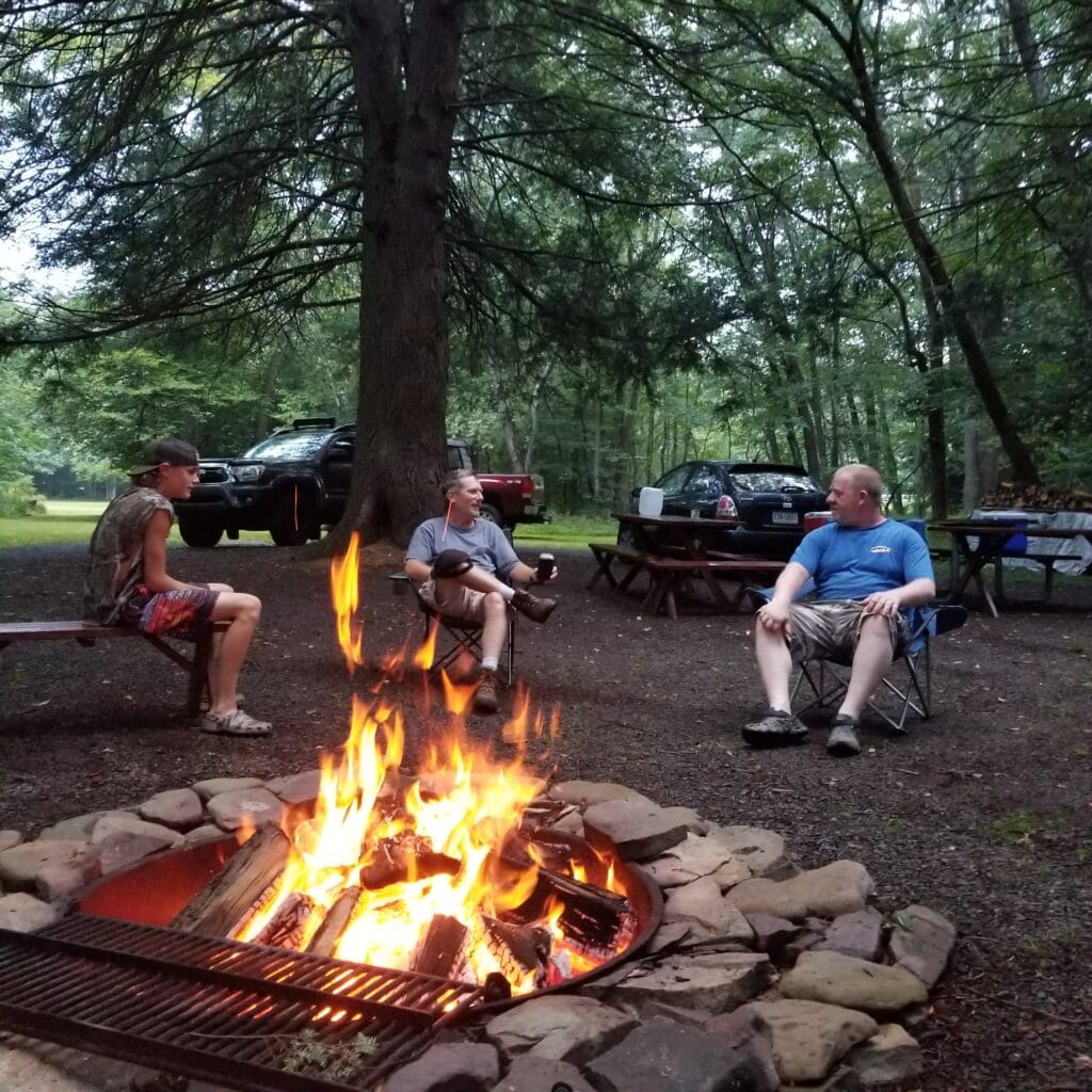 3 guys sitting in camp chairs around a blazing fire in a stone firepit underneath trees talking