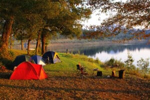 campsite alongside a lake with 3 tents under trees and chairs around a firepit